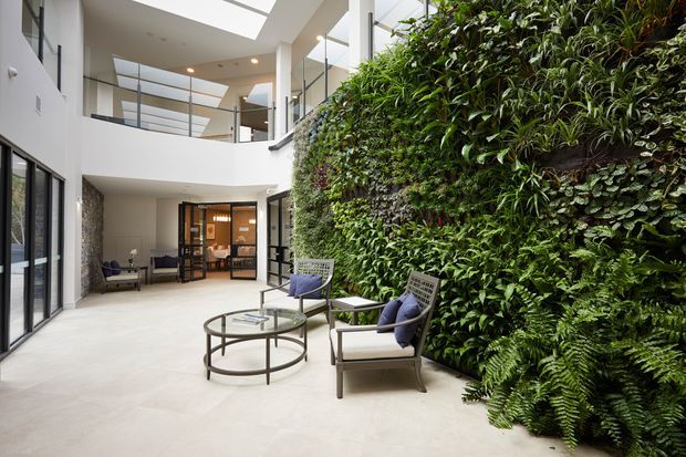 A bright, contemporary multi-level interior courtyard or atrium with a large living green wall, skylights, glass balustrades, pale flooring, and a small lounge arrangement of armchairs and a round coffee table. An enclosed meeting or dining area is visible beyond the seating.