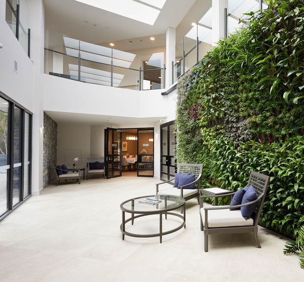 A bright, contemporary multi-level interior courtyard with a large living green wall, pale tiled flooring, glass balustrades, skylights, and floor-to-ceiling glazing. Outdoor-style lounge chairs and a circular glass coffee table create a refined seating area, while a dining space is visible beyond the glazed doors. The space is filled with natural light and features a double-height void.