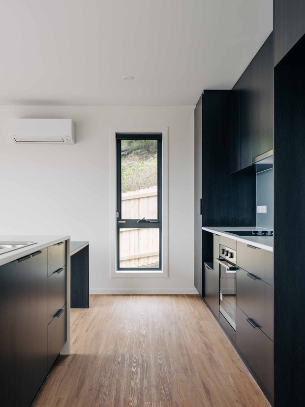 A contemporary galley kitchen viewed lengthwise, with dark charcoal built-in cabinetry on both sides, pale stone or composite benchtops, integrated stainless-steel appliances, and warm light timber flooring. White walls and ceiling create a bright, restrained interior, with a narrow full-height window at the end and a wall-mounted split-system air conditioner.