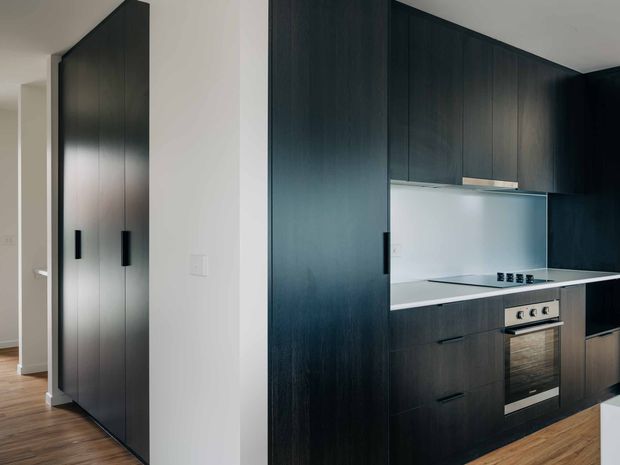 A compact contemporary kitchen with tall dark timber-look cabinetry, integrated black handles, a pale benchtop, and a light reflective splashback. The cabinetry extends into a full-height storage wall beside the kitchen, while timber flooring continues through the open-plan space. A stainless-steel oven and cooktop are built into the lower joinery.