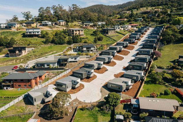 Aerial view of a newly developed residential hillside subdivision, with rows of compact modern detached homes arranged along a broad concrete access road. The houses have dark grey roofs, contemporary rectangular forms, and landscaped front gardens, set among established homes and heavily vegetated sloping terrain.