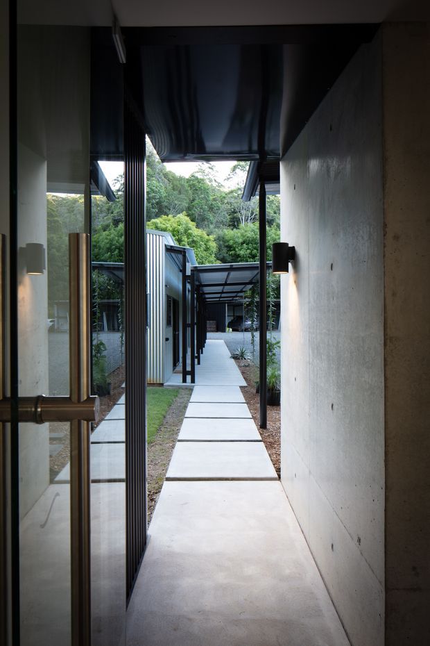 A narrow covered passage leading from a glazed doorway through a landscaped residential courtyard. The scene features smooth concrete walls and paving, dark slender metal posts and roof soffits, extensive glazing, and a straight path of large rectangular concrete slabs bordered by gravel, lawn, and dense greenery. The image has a restrained, modern architectural character with strong indoor-outdoor connection.