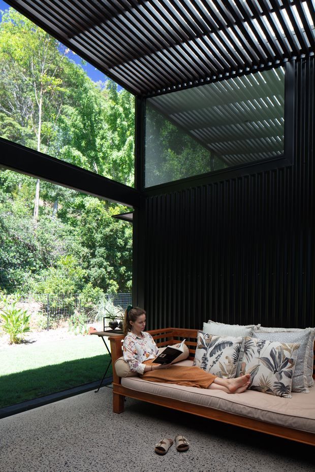 A sunlit indoor-outdoor lounge enclosed by large glass walls, with a timber-framed sofa, patterned cushions, and a woman reading. The space features dramatic black vertical wall cladding, expansive glazing, and a dark slatted pergola or louvred ceiling that filters sunlight. A leafy garden is visible outside, creating a strong connection to nature.
