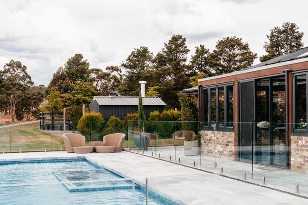 Exterior view of a contemporary brick-and-glass house beside a swimming pool. The terrace has frameless glass balustrades, pale paving, and curved outdoor lounge chairs, with a dark metal-clad outbuilding and landscaped property beyond. The building features large black-framed glazed openings and a low-pitched metal roof.