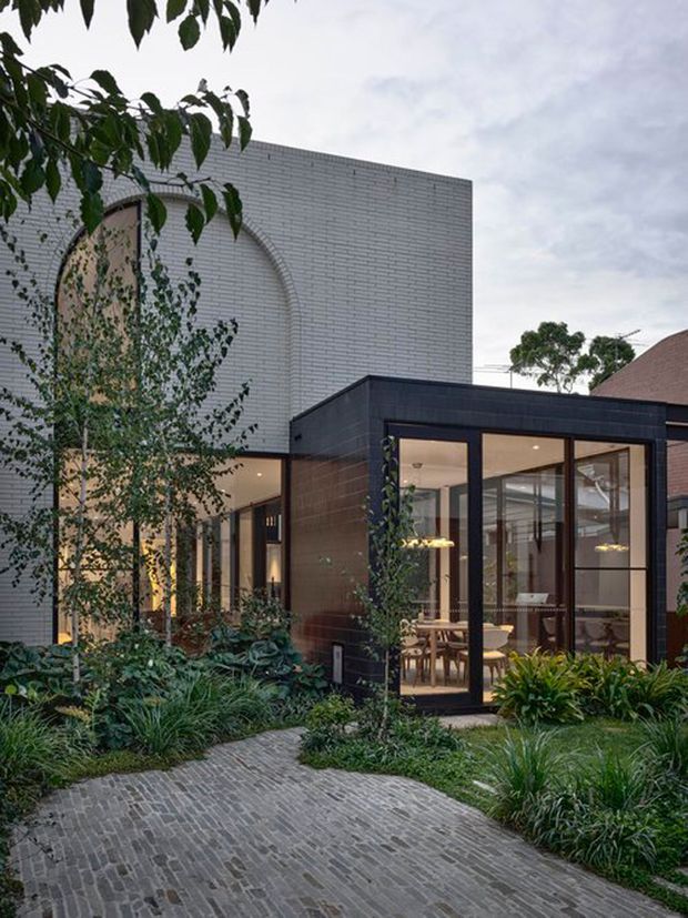 Exterior view of a contemporary residential extension framed by lush planting. The composition features a tall light-grey brick volume with a large arched opening, a darker brick-and-glass pavilion with extensive floor-to-ceiling glazing, and a curved grey brick-paved path leading through the garden. Warm interior lighting and a dining area are visible through the windows.