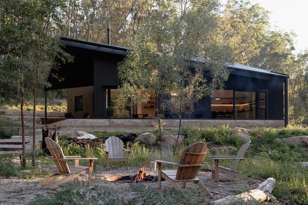 A low, modern single-storey pavilion-style home set within a wooded landscape. The dark vertically ribbed exterior has broad floor-to-ceiling glazing, projecting flat roof planes, and warm timber interior finishes visible through the windows. In the foreground, timber Adirondack chairs surround an outdoor fire pit on a gravel terrace, with planted landscaping and exposed rocks around the building.