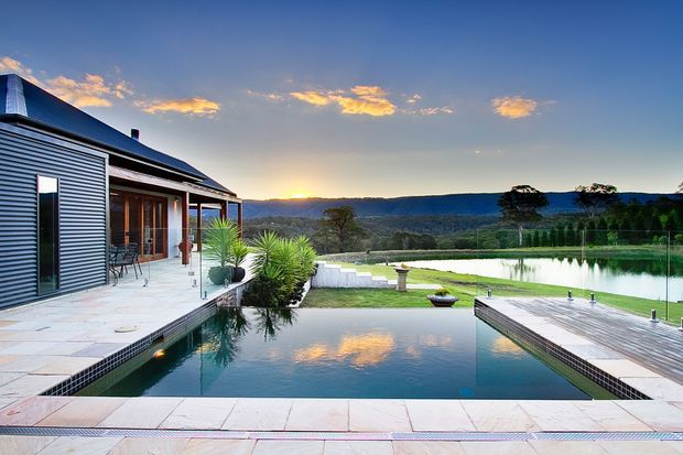 Contemporary single-level home with corrugated metal cladding, extensive glazing, and a broad paved terrace overlooking a rectangular swimming pool. The landscaped property includes a timber pool deck, clipped hedges, ornamental planting, and expansive views toward a wooded, elevated landscape at sunset.