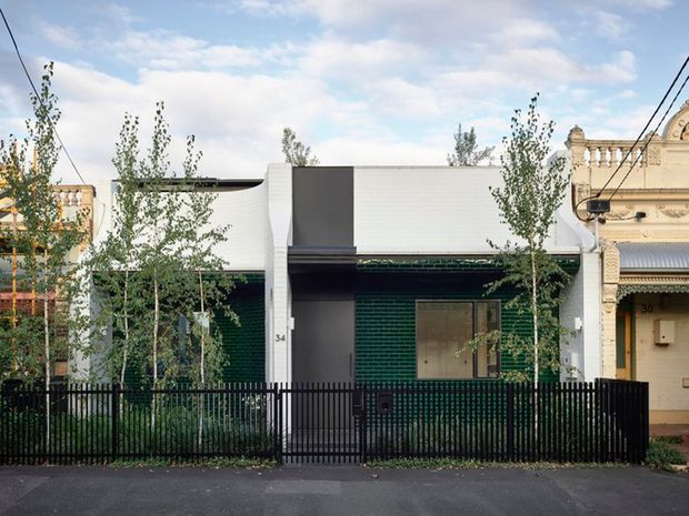 Street-facing view of a compact contemporary infill dwelling between ornate heritage terrace-style facades. The home has a dark green glazed-brick facade, crisp white horizontal upper cladding, charcoal panels, a simple flat roofline, a small front window, and a black metal picket fence. Young street trees partially screen the frontage.