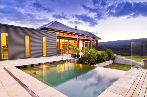 Twilight exterior view of a modern hillside home with dark horizontal cladding, a prominent pitched metal roof, warm-lit glazed living areas, and a tiled swimming pool terrace. Tropical-style planting surrounds the pool, with expansive distant hills visible beyond the property.