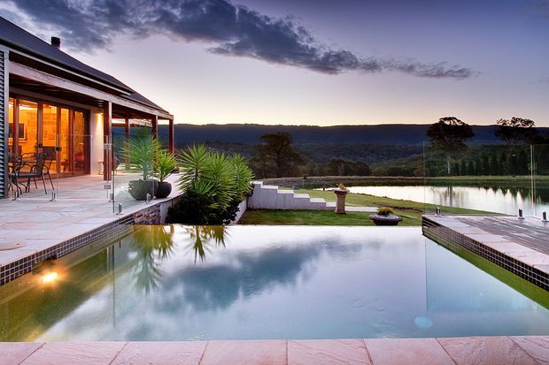 A dusk exterior view of a modern single-storey residence beside a large reflective swimming pool. The home has dark pitched roofing, extensive glazed doors, a covered outdoor terrace with dining furniture, pale tiled paving, and landscaped architectural planting. Beyond the pool is a broad rural or bushland outlook, with warm interior lighting creating an inviting atmosphere.