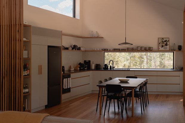 Warm contemporary open-plan kitchen and dining area with a high ceiling, clerestory window, timber flooring, light built-in cabinetry, integrated dark refrigerator, tiled backsplash, and a large horizontal window overlooking trees. A timber slatted screen and open shelving add vertical texture, while a pendant light hangs above the dining table.