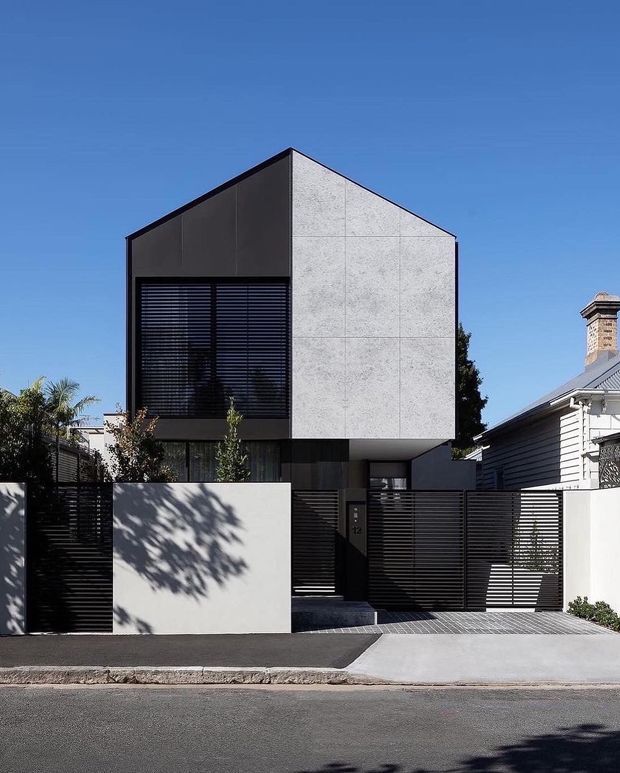 Street-facing exterior photograph of a contemporary two-storey residence with a sharply pitched, gabled form. The upper facade combines dark charcoal cladding with large pale textured panel cladding, while a broad window is screened by horizontal dark louvres. The lower level is recessed behind dark horizontal slat fencing and a pale rendered boundary wall, with a concrete driveway and pedestrian entry. A neighbouring weatherboard house is visible at right.