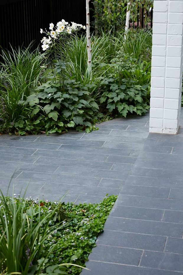 A landscaped courtyard or garden walkway paved with dark grey rectangular stone tiles in a staggered layout. Dense ornamental planting, grasses, flowering plants and a slender tree border the paving, while a white-painted brick column frames the right side beside a dark recessed structure.