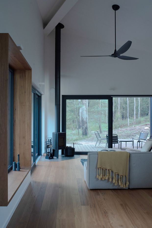 A calm, modern living room with a tall white ceiling, pale timber flooring, built-in timber cabinetry, and a black wood-burning stove with a vertical flue. A large black-framed glazed opening connects to a timber deck and wooded landscape outside. A contemporary sofa, ceiling fan, and simple outdoor furniture are visible.