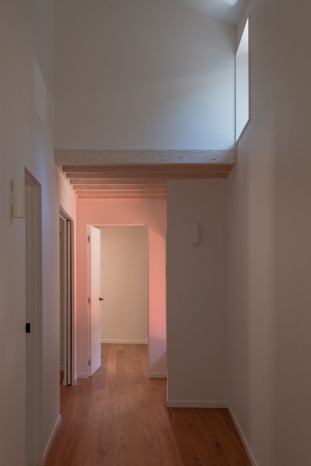 A narrow residential hallway viewed toward an open doorway, with warm timber flooring, white painted walls, and a timber-lined overhead void or landing. Exposed light timber beams span the corridor near the ceiling, while soft pink and cool daylight create a subdued atmospheric glow. Several doors and a slim window opening are visible, emphasizing the vertical circulation space.