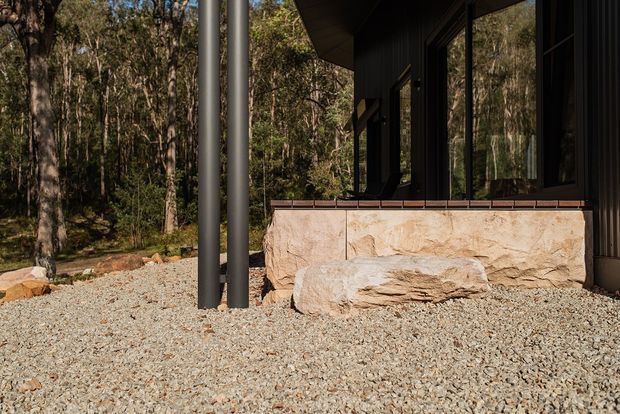 Exterior detail of a dark-clad contemporary building set within a wooded landscape. The view focuses on a raised stone-clad base, black-framed glazing, slender dark structural posts, and a pale gravel ground plane, with dense trees surrounding the structure.