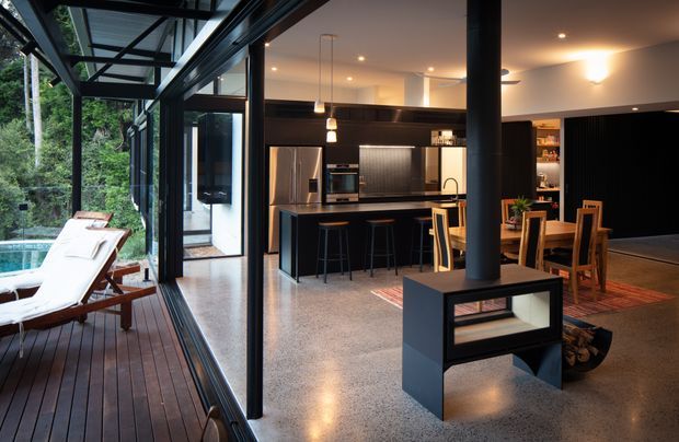 Open-plan contemporary kitchen, dining, and living area viewed from a glazed indoor-outdoor connection. The space features extensive black-framed glazing, a dark kitchen with island seating, stainless-steel appliances, polished concrete or terrazzo-like flooring, timber dining furniture, and a freestanding black wood-burning fireplace. A timber deck and leafy outdoor setting are visible beyond the glazing.
