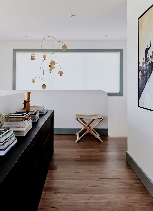 A refined contemporary interior, likely a living room or sitting area, with warm timber flooring, white walls, a large recessed frosted or translucent window, and a sculptural brass pendant light. A curved white partition or built-in seating element frames a small folding stool. In the foreground, a dark built-in cabinet is topped with stacked books and a timber decorative object, while framed artwork adds visual interest on the right wall.