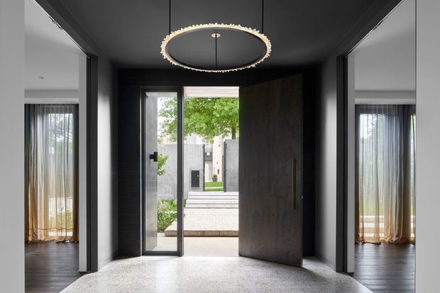 A dramatic modern entry foyer with a dark, charcoal-toned ceiling and walls, pale textured flooring, and a large circular pendant light. A tall pivot door and glazed side panels open toward a landscaped courtyard, while adjacent rooms with sheer curtains are visible on both sides.