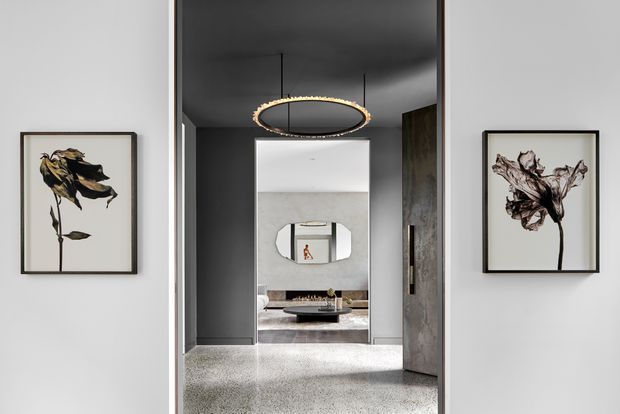 A refined contemporary interior view through a wide hallway or entry passage, with dark charcoal walls and ceiling, pale terrazzo flooring, framed botanical artwork, and a sculptural suspended oval pendant light. An open doorway reveals a softly furnished living area with a concrete-toned feature wall and rounded mirror.