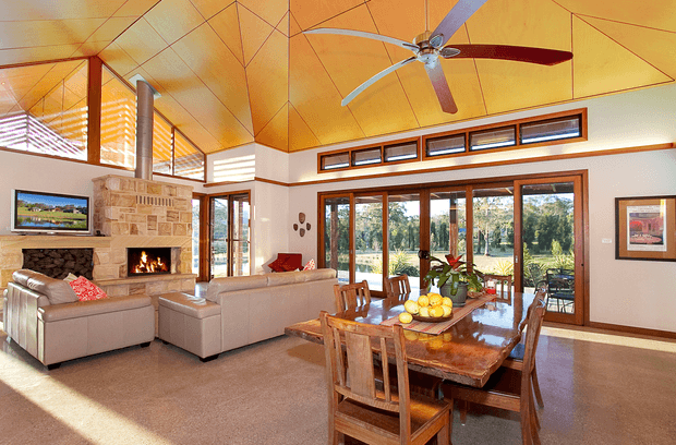 Bright open-plan living and dining room with a high angular timber-lined ceiling, exposed geometric beams, a large ceiling fan, and extensive timber-framed glazing overlooking greenery. The living area includes a stone-clad fireplace with a lit fire and neutral upholstered sofas, while the dining area has a substantial polished timber table and chairs. Warm natural light, timber detailing, and indoor-outdoor visual connection create an inviting contemporary interior.
