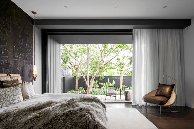 A refined contemporary bedroom with a low-profile bed dressed in layered neutral linens and a plush grey throw. The room features a dark textured stone feature wall, suspended pendant lighting, charcoal structural beam, sheer floor-to-ceiling curtains, and large black-framed glazing opening to a planted outdoor terrace. A sculptural leather lounge chair sits beside the window, creating a calm indoor-outdoor connection.