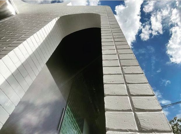 Low-angle exterior view of a tall white-painted masonry facade with a deep arched recessed entry or opening. The facade has a regular block or brick pattern and a small section of green-tinted glazing near the base. Bright blue sky and clouds are visible around the structure.