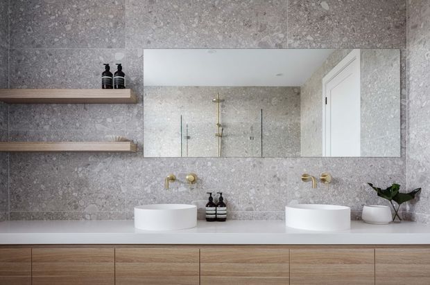 Contemporary double-vanity bathroom with a long light-wood floating vanity, two white vessel basins, wall-mounted brushed-gold taps, and a large horizontal mirror. Grey terrazzo-style wall tiles continue around a glass shower enclosure, with simple timber display shelves and minimal accessories.