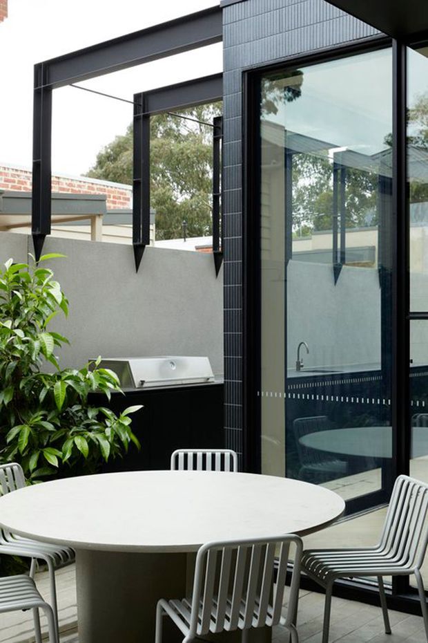 A contemporary courtyard or outdoor dining terrace with a large round light-coloured table and white slatted chairs. The space includes a built-in black barbecue beneath a minimalist steel pergola, a rendered boundary wall, dark tiled exterior cladding, and large black-framed glazed doors reflecting the courtyard. Green planting softens the otherwise restrained monochrome palette.