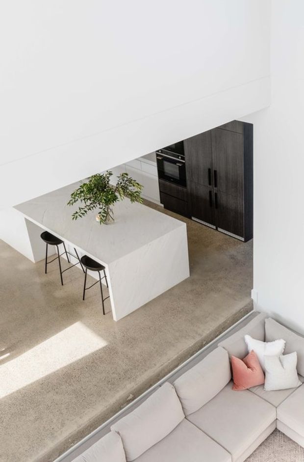 High-angle view of a bright, minimalist open-plan kitchen and living area. A large white waterfall island with seating for three occupies the kitchen, paired with dark wood-grain full-height cabinetry and integrated black appliances. The space has a polished concrete floor, white walls and ceiling, and a pale sectional sofa with soft cushions in the foreground. Strong natural light creates a clean, airy atmosphere.