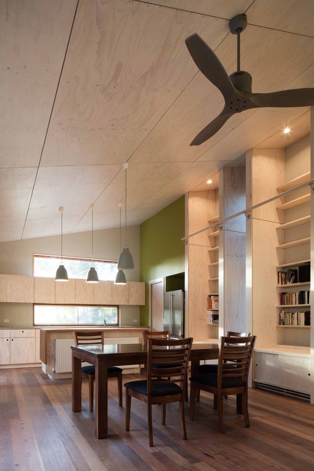 Warm, compact open-plan kitchen and dining area with extensive pale plywood wall cabinetry and built-in shelving. A dark timber dining table and chairs sit beneath pendant lights, while a ceiling fan hangs from the plywood-lined sloping ceiling. The room features a muted green accent wall, timber flooring, stainless-steel refrigerator, and integrated radiator and storage.