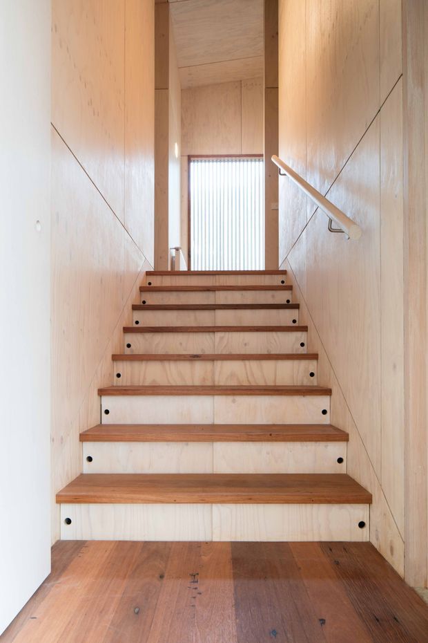 A narrow, straight-run timber staircase enclosed by pale plywood wall and ceiling panels. The stair treads and landing are darker warm timber, with exposed black fasteners on the stair risers and a simple timber handrail on the right. A tall narrow window with vertical screening provides soft daylight at the upper landing, creating a warm, minimalist interior.