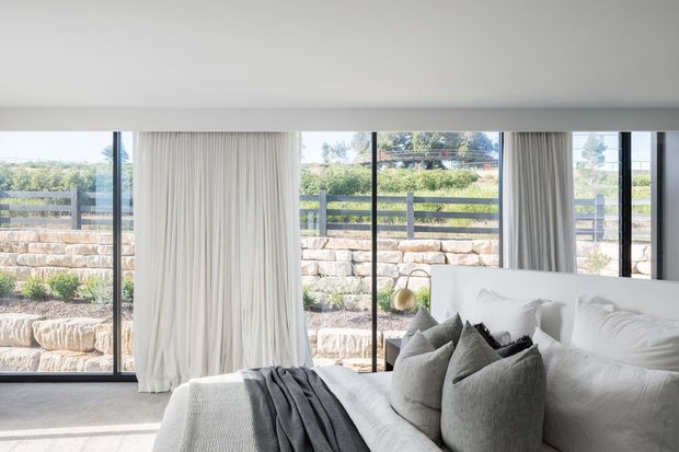 Bright contemporary bedroom interior with a large white bed, layered grey and white cushions, a draped charcoal throw, floor-to-ceiling glazing, and full-height sheer curtains. The room opens visually to a landscaped stone terrace and garden beyond.