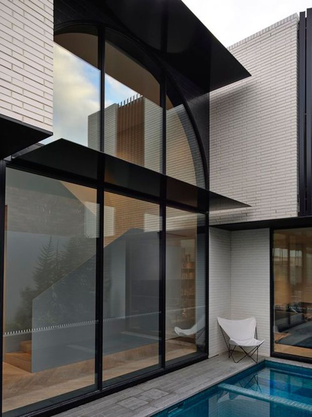 A contemporary multi-level courtyard facade with crisp white brick walls, extensive black-framed glazing, dramatic projecting black roof and canopy forms, and a small swimming pool along the paved terrace. Interior spaces and a sculptural white lounge chair are visible through the glass.