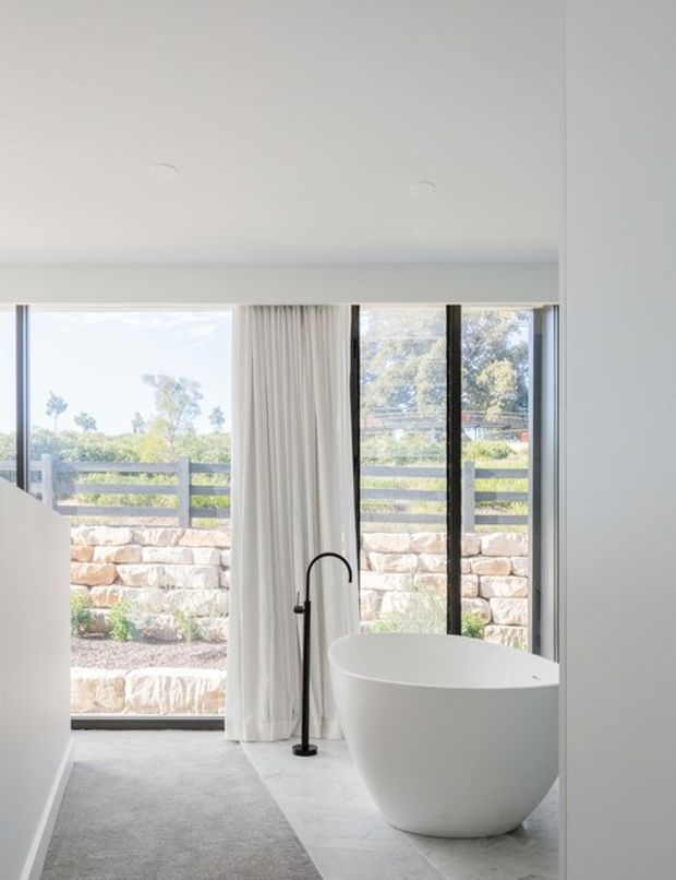 Bright minimalist bathroom featuring a freestanding white oval bathtub beside full-height black-framed glazing, a black floor-mounted bath filler, pale stone or porcelain floor tiles, soft white curtains, and a grey rug. The glazing overlooks a landscaped stone retaining wall and garden.