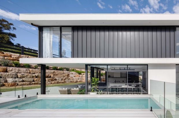 Exterior view of a modern two-storey residence with a flat roof, crisp white horizontal slabs, dark vertical metal cladding, and extensive black-framed glazing. The lower level opens directly onto a swimming pool and terrace, with stepped sandstone retaining walls and landscaped ground around the house. A contemporary kitchen and dining area are visible through the glass.