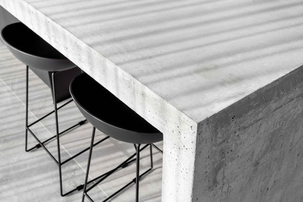 Close-up architectural detail of a thick, raw concrete kitchen island or counter with a ribbed surface, paired with two black metal-framed bar stools. The monochromatic composition emphasizes brutalist materiality, strong geometric planes, and textured concrete.