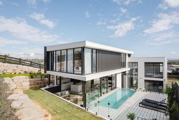 A contemporary multi-level house on a landscaped sloping site, featuring a crisp white and dark-framed facade, extensive floor-to-ceiling glazing, a projecting upper-level box, and a rectangular swimming pool beside a timber-toned deck. Stone retaining walls and stepped planting frame the property, with broad suburban views beyond.