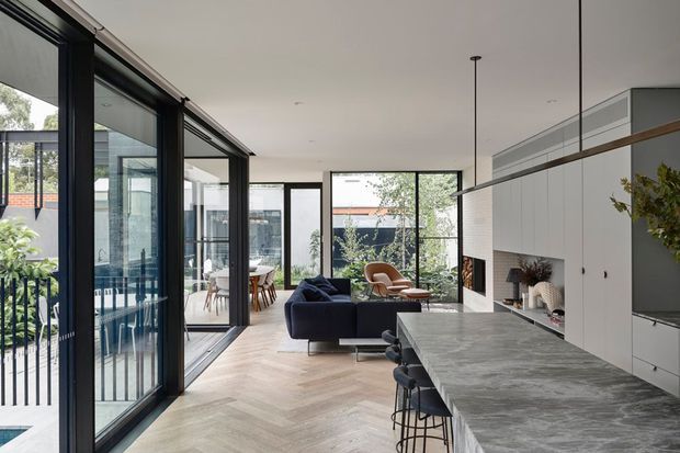 A bright, contemporary open-plan interior combining a kitchen, dining area and living room. The space features a large grey stone island with dark stools, white built-in cabinetry, a navy sofa, timber herringbone flooring, black-framed floor-to-ceiling glazing and a linear pendant light. Indoor planting and views to an exterior courtyard add a calm, airy quality.