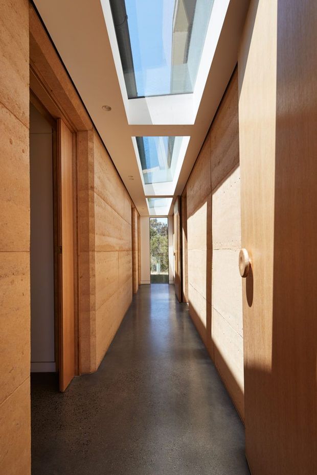 A long, narrow interior hallway with warm timber door and trim detailing, exposed horizontal-banded stone or rendered masonry walls, a dark polished concrete floor, and a series of rectangular skylights bringing strong natural light into the corridor. Open doorways line the hall, with glazing and trees visible at the far end.