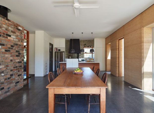 Open-plan dining and kitchen interior with a long solid timber dining table and chairs in the foreground. The room combines exposed red brickwork on the left, warm timber wall panelling and doors on the right, white cabinetry, dark polished concrete flooring, and a compact kitchen with a dark suspended rangehood. Large windows and glazing provide strong natural light, while a white ceiling fan hangs overhead.