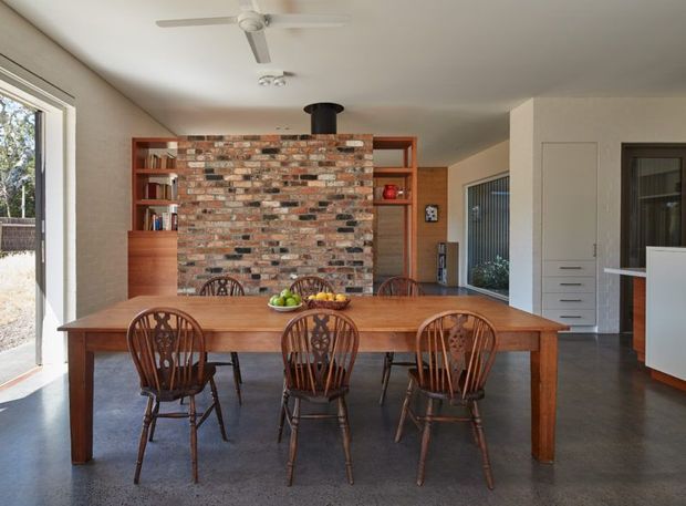 A bright open-plan dining area with a long solid timber dining table and ornate wooden chairs. A red-toned exposed brick feature wall with integrated timber shelving and cabinetry forms the backdrop, with a black cylindrical fireplace flue above. The room has polished grey concrete flooring, white plaster walls and ceiling, large glazing on the left, built-in white cabinetry on the right, and a ceiling fan. The overall character is warm, earthy and mid-century influenced.