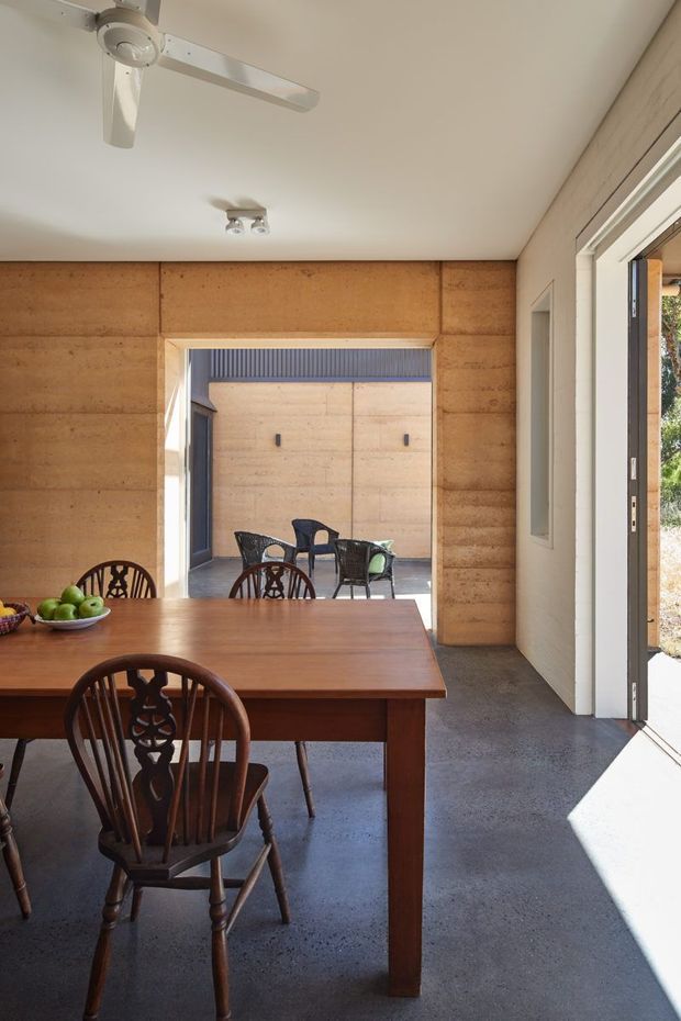 A bright dining room with a long solid timber dining table and traditional spindle-back chairs. Warm timber wall panelling surrounds a wide opening to a small paved courtyard, while a large glazed door brings in strong sunlight. The room has a smooth white ceiling with a ceiling fan and compact spotlight, and a dark polished-looking concrete floor. The interior combines warm natural timber with simple, functional architectural detailing.