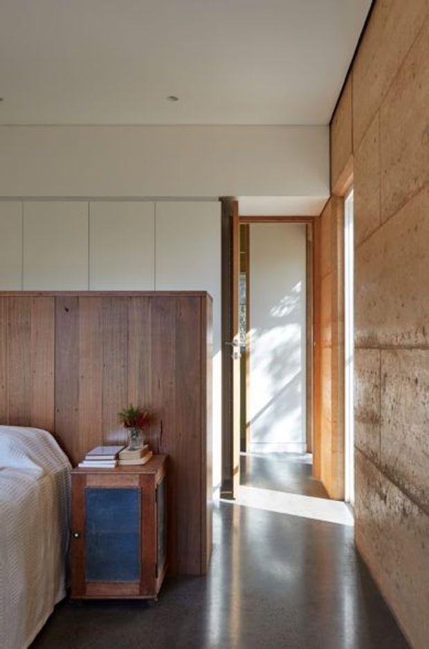 A warm, minimalist bedroom viewed toward an open doorway and adjacent hall. The room features a partially visible upholstered bed, a compact dark timber bedside cabinet, a full-height timber headboard or storage wall, smooth pale walls and ceiling, polished dark concrete flooring, and a textured earthen or rammed-earth feature wall along the right side. Natural light enters through the doorway and narrow glazing, creating a calm, earthy atmosphere.