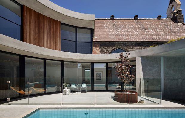 A contemporary curved courtyard addition wraps around a swimming pool, combining smooth concrete bands, dark-framed floor-to-ceiling glazing, vertical timber cladding, and a glazed corner. A weathered brick building with a pitched tiled roof and chimney forms the contrasting heritage backdrop. The courtyard includes a central raised planter and minimalist outdoor seating.
