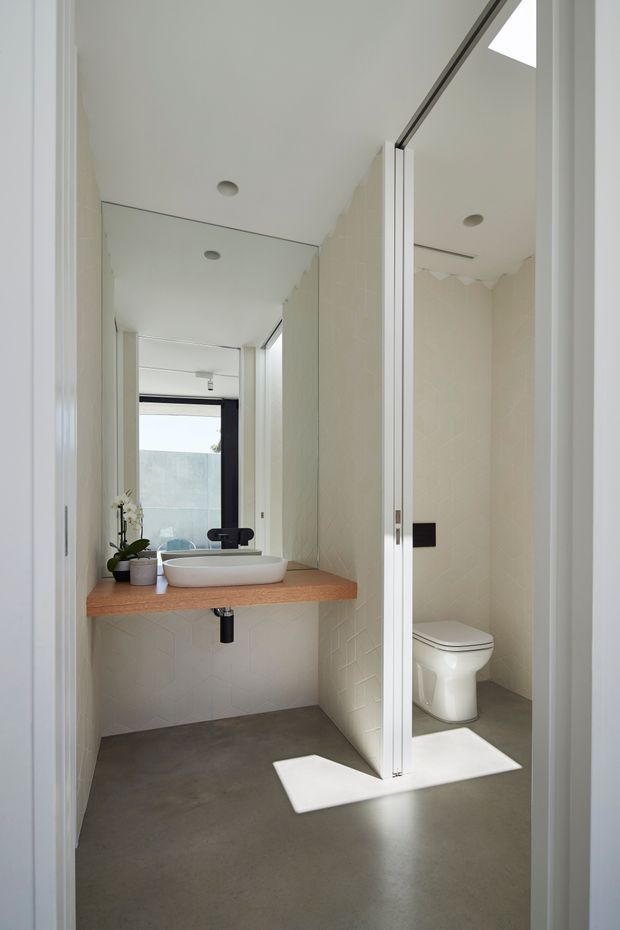 Bright contemporary bathroom viewed through its doorway, featuring a floating timber vanity with a white vessel basin, large mirrored wall, concealed wall-mounted tap, white toilet in a partially screened alcove, pale geometric wall tiles, polished concrete floor, recessed downlights, and a skylight. Natural light creates a clean, minimalist atmosphere.
