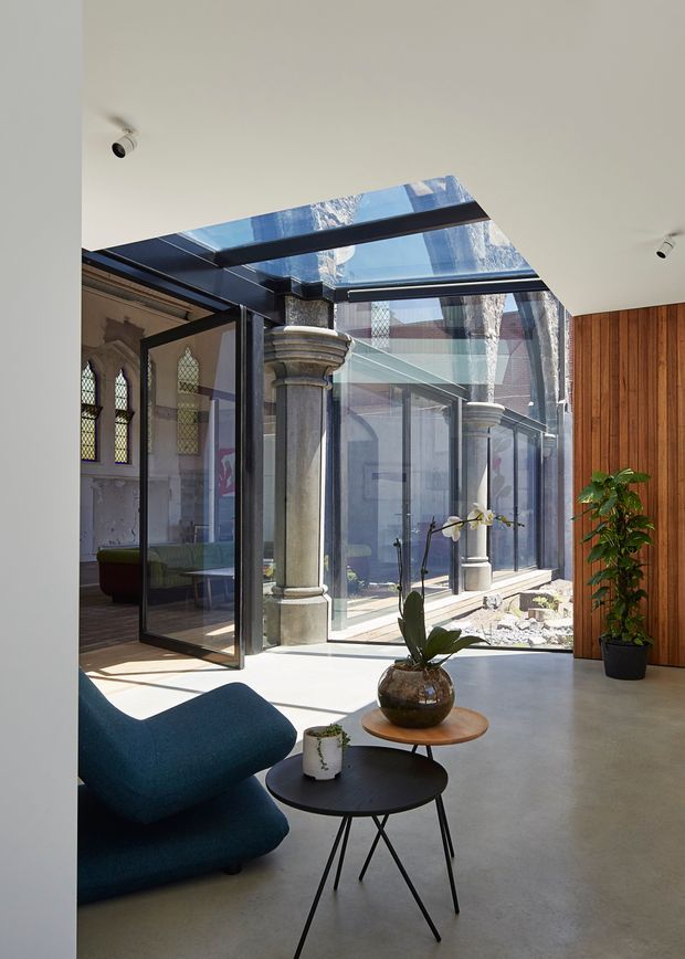A bright contemporary interior courtyard or sunroom with a glazed roof and full-height glass walls enclosing a landscaped atrium. Classical stone columns and arched windows from an adjoining heritage structure contrast with dark metal glazing frames, pale concrete flooring, vertical timber wall panelling, and modern blue lounge seating. Potted plants and small round tables furnish the space.