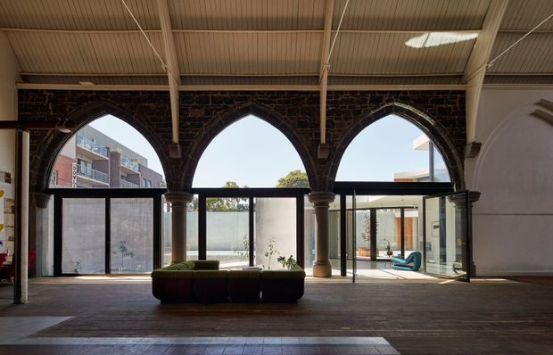A dramatic covered interior courtyard or atrium with three tall pointed Gothic-style arches in dark exposed brick, supported by stone or concrete columns. Large glazed openings and sliding glass doors connect to an outdoor courtyard, while a corrugated metal canopy with exposed structural beams spans overhead. The space has dark timber or wood-look flooring, a central low upholstered bench, and strong natural daylight.