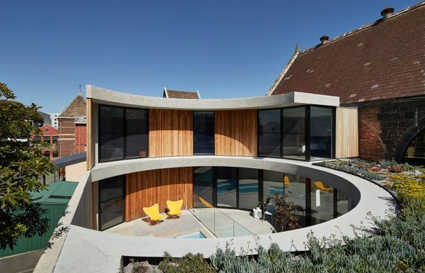 Exterior view of a contemporary curved two-storey addition wrapped around a landscaped courtyard. The addition features extensive dark-framed glazing, vertical timber cladding, sweeping concrete slab edges and a curved roof terrace planted with succulents. It is attached to a retained historic brick and stone building with a steep tiled roof, creating a clear contrast between old and new architecture.
