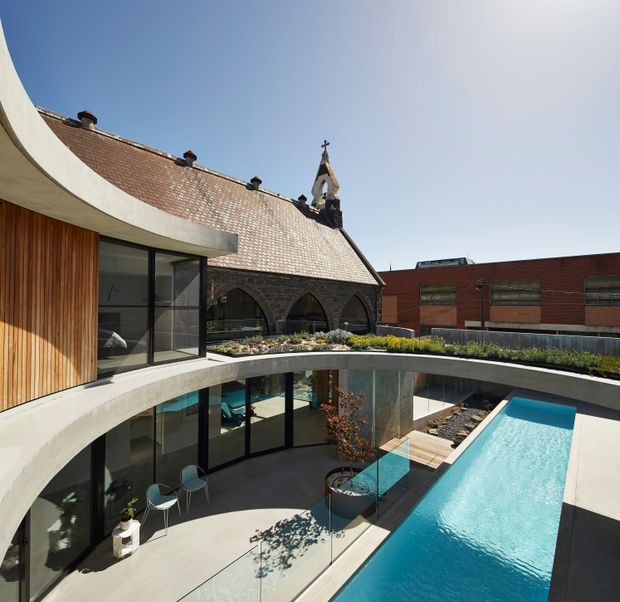 A contemporary multi-level courtyard building wraps around a long turquoise swimming pool. The foreground structure has sweeping curved concrete balconies, warm vertical timber cladding, extensive black-framed glazing and a glass balustrade. Beyond it is a preserved masonry church-like building with a steep tiled roof, arched openings and a small bell tower, alongside a modern red-brick structure. Bright sunlight and clear sky create a strong contrast between the historic and contemporary architecture.